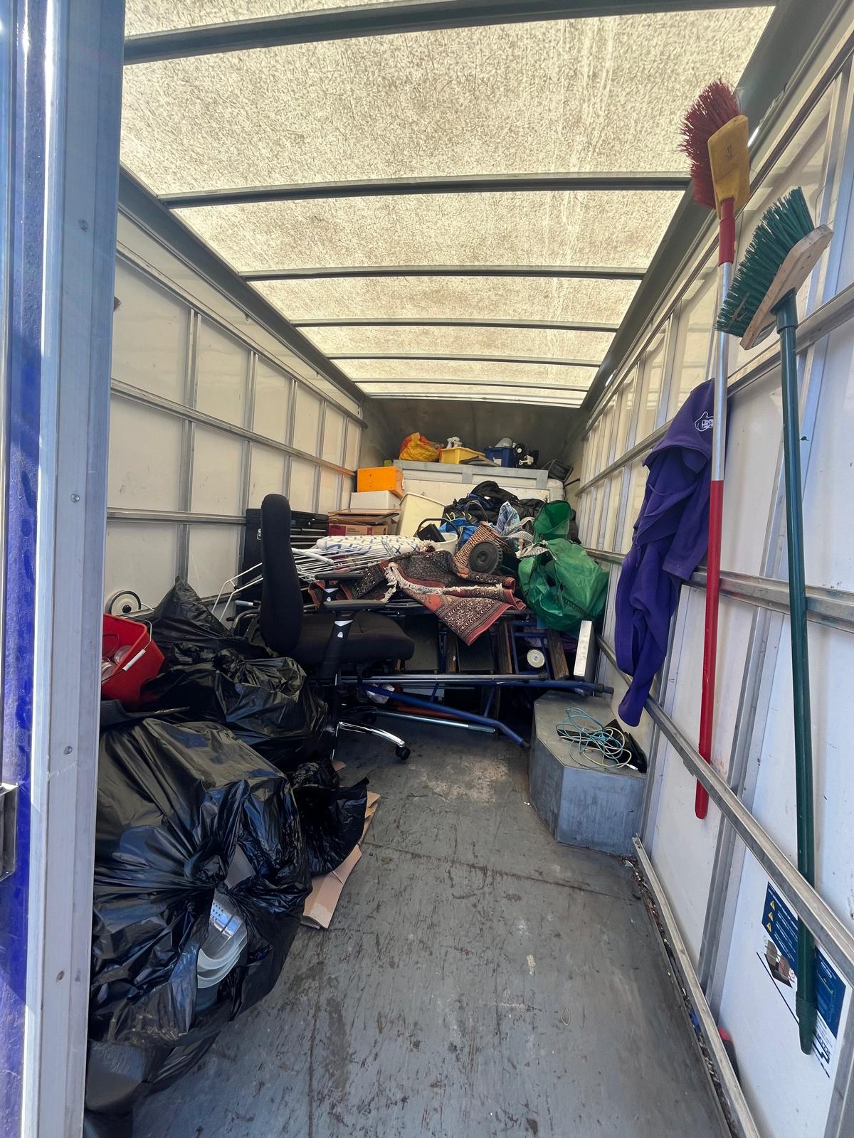 Inside a loaded clearance van with black bags, an office chair, rugs, metal frames and boxes stacked to the roof