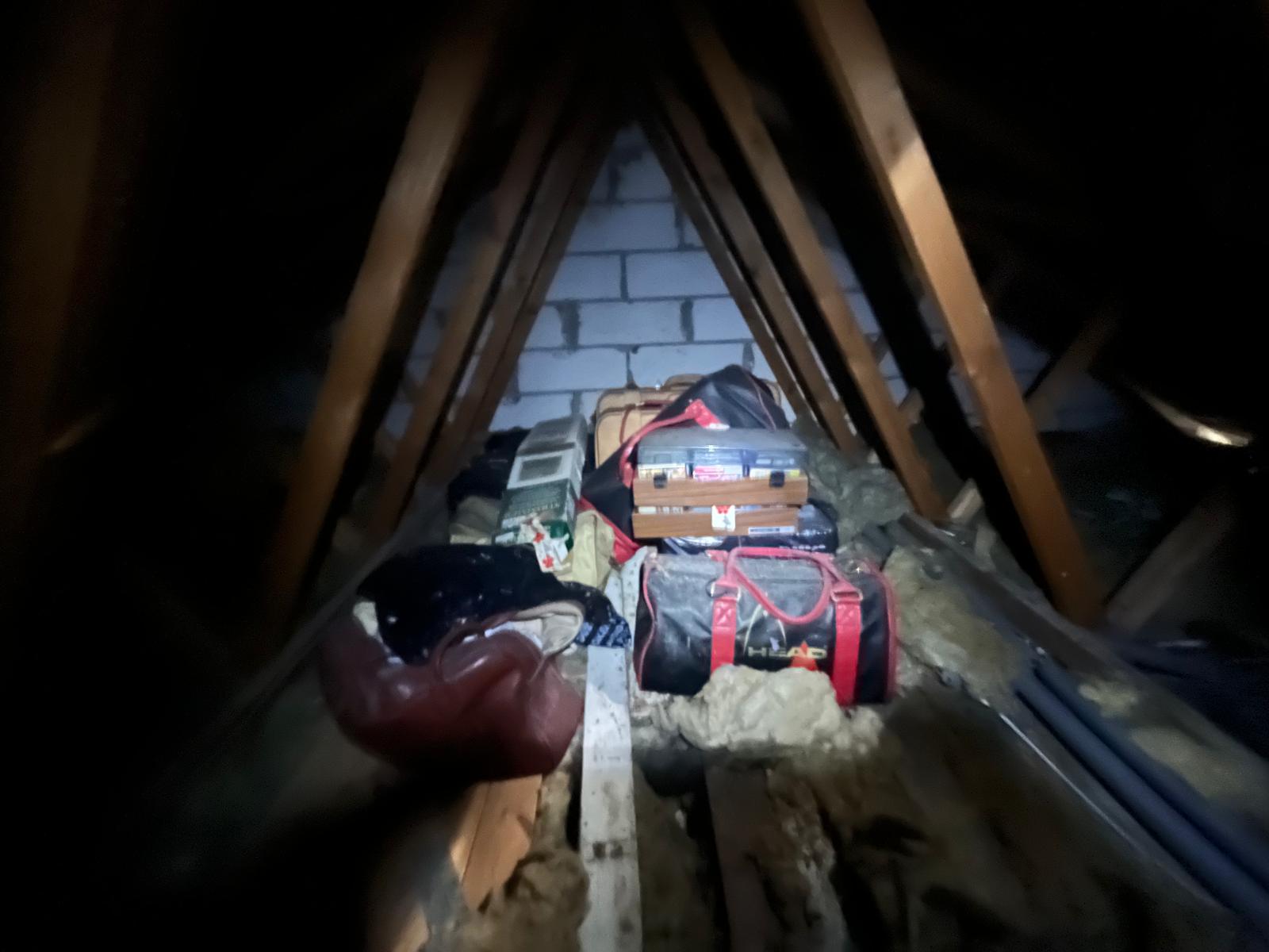 Loft interior lit by torchlight showing holdalls, hard suitcases, an old stereo and bags stacked on top of the insulation between the joists