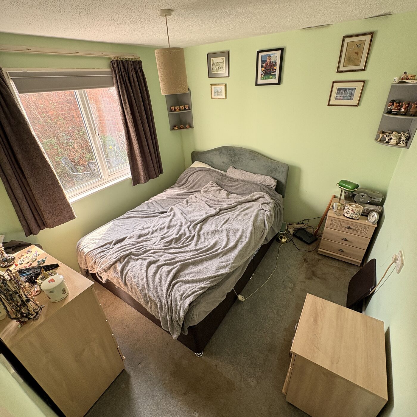 Green bedroom with a made-up double divan bed, bedside cabinet, two chests of drawers and framed pictures and ornaments on the walls